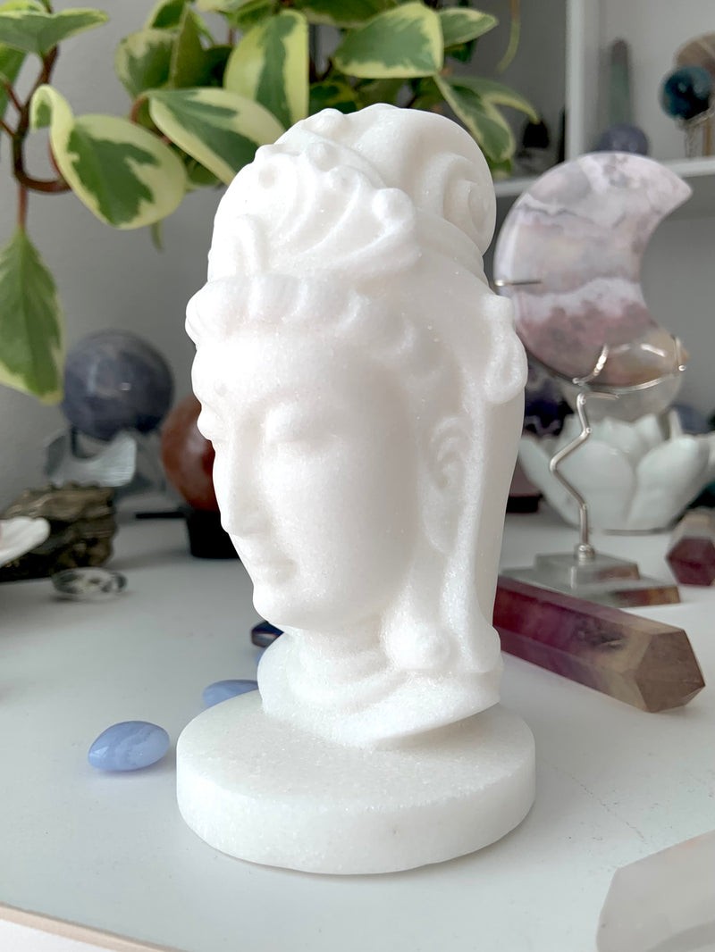 White Jade Quan Yin carving shown on a white table with other crystals and a plant in the background