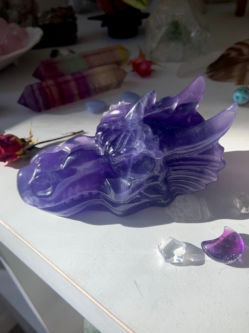 Large purple fluorite crystal dragon head carving shown on a white table with other crystals in the background