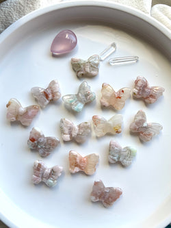 An array of Mini pastel Flower Agate Butterfly Carvings shown on a white plate with some other small crystals