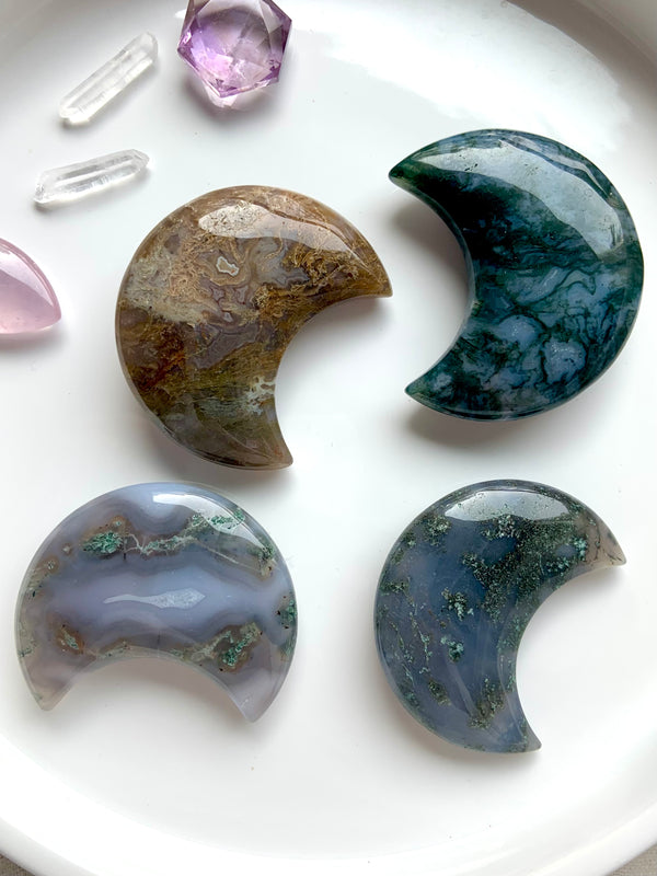 Colorful Moss Agate Moons shown on a white place with some other small crystals in the background. These moons are a chunky shape that is nice to hold like a palm stone in meditation. Choose your favorite!