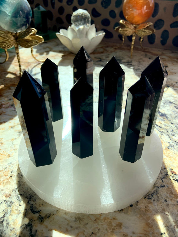 An array of black obsidian points shown in a group on top of a selenite plate with crystal spheres in the background. Choose your size from the dropdown menu.